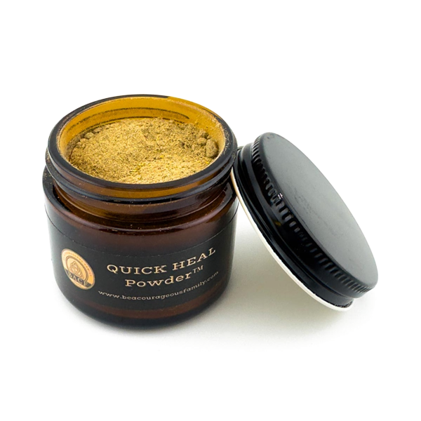 An open amber glass jar of Quick Heal Powders showing the golden-brown powder inside, with its black lid leaning against it on a white background.