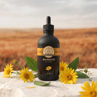 A 4 ounce black dropper bottle of Arnica tincture set on a white stone, surrounded by arnica flowers with a warm desert prairie background.